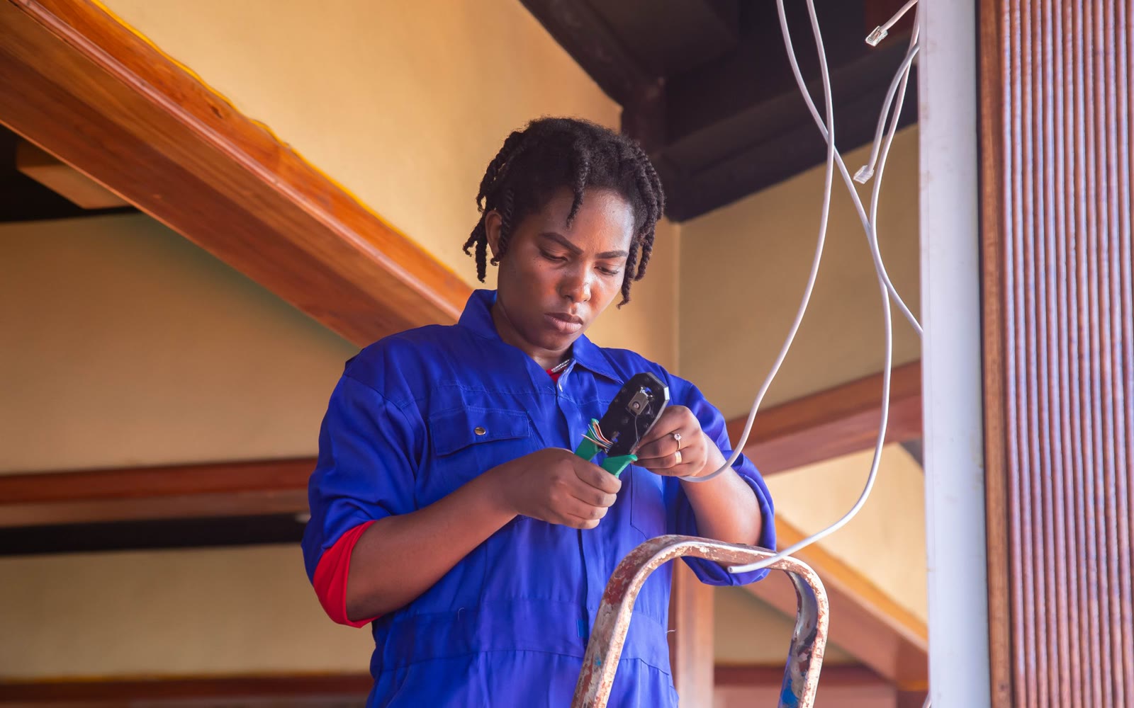 An electrician in blue coveralls stripping a wire on a ladder