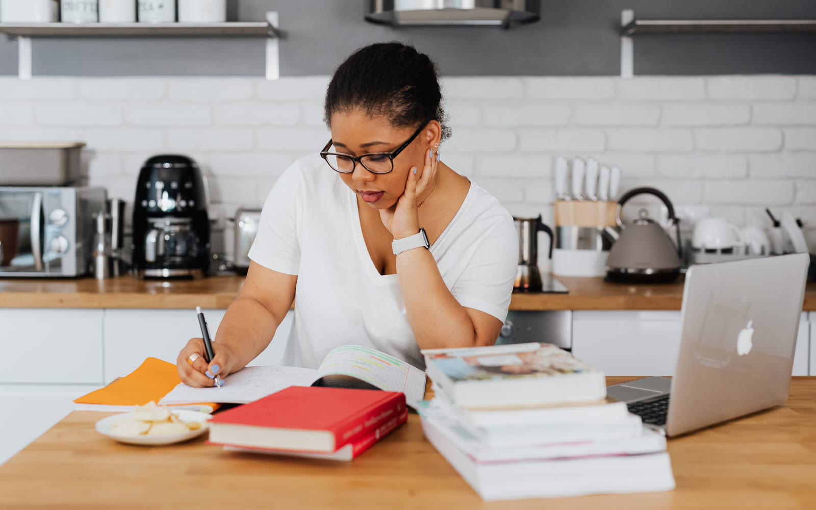 A woman studying at her kitchen table with textbooks, a notebook and a laptop